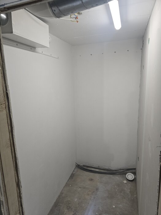 Rum med vitmålade väggar, synliga ventilationsrör, LED-belysning och betonggolv. Dåligt förberett för målning och renovering.