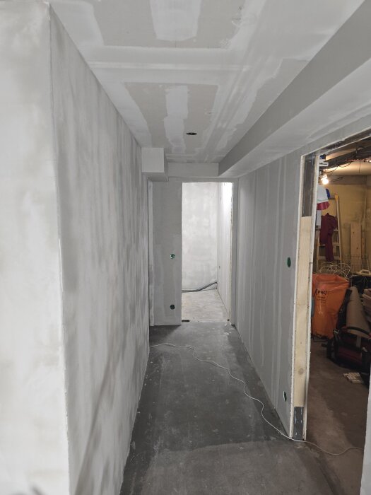 Gång i källaren med nymonterade gipsskivor och spacklade tak; en byggarbetsplats med synliga kablar och öppningar mot intilliggande rum.