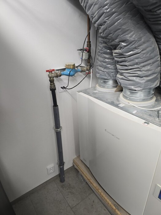 Vattenmätare och gråa ventilationsrör installerade i ett maskinrum med klinkergolv.