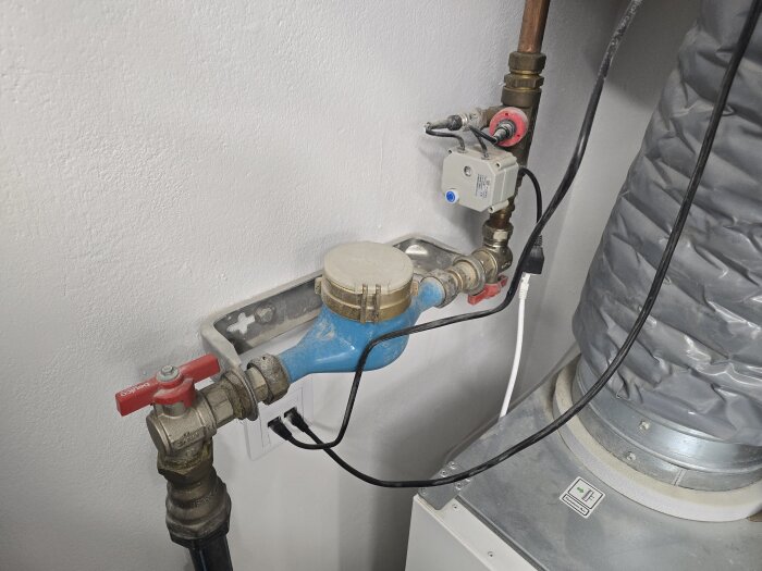 Vattenmätare med blå kropp och tillhörande rörsystem, omgiven av ventilationsutrustning, installerad mot en vit vägg.
