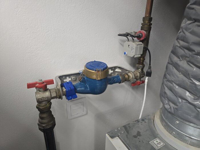 Blå och gul vattenmätare installerad på en vit vägg med rör och ventiler, till höger syns en grå slang.