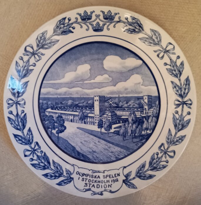 Tallrik med motiv av Stockholms stadion under Olympiska spelen 1912, dekorerad med löv och kronor.