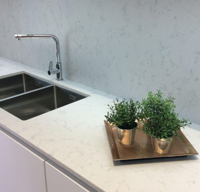 Köksbänk i kompositsten med integrerad diskho och en vägg i liknande material, dekorerad med växter i krukor på en bricka.