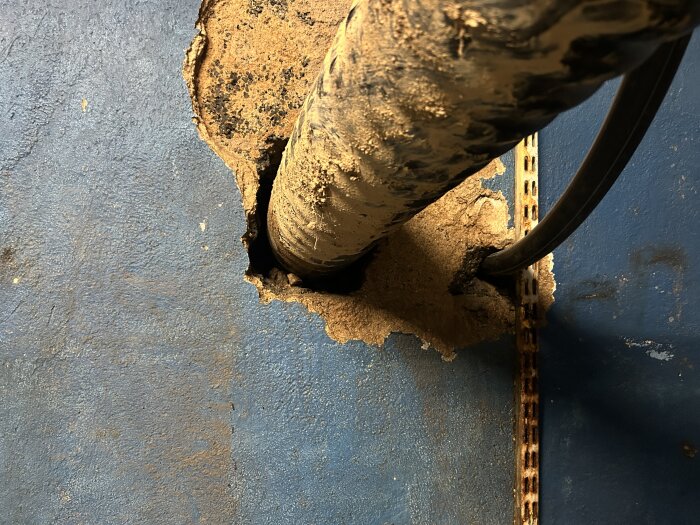 Hål i blått vägg där en tjock kulvert passerar, omger med sprucket murbruk och rostig metallremsa, visar otäthet och problem med vatteninträngning.