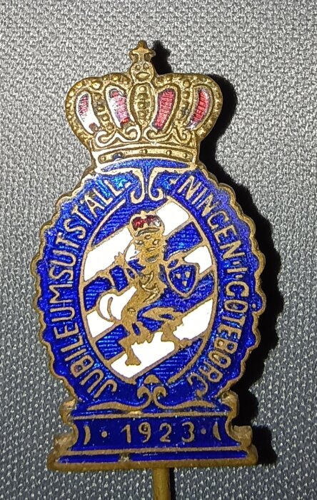 Nålmärke från Göteborgsutställningen 1923 med kungakrona, blå lejonemblem och texten "Jubileumsutställningen Göteborg".