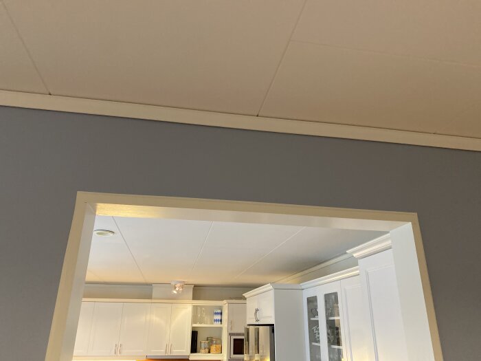 Öppning mellan kök och hall utan foder, mdf-skivor användes för att ersätta rundat valv. Kök med vita skåp och grå väggar syns i bakgrunden.