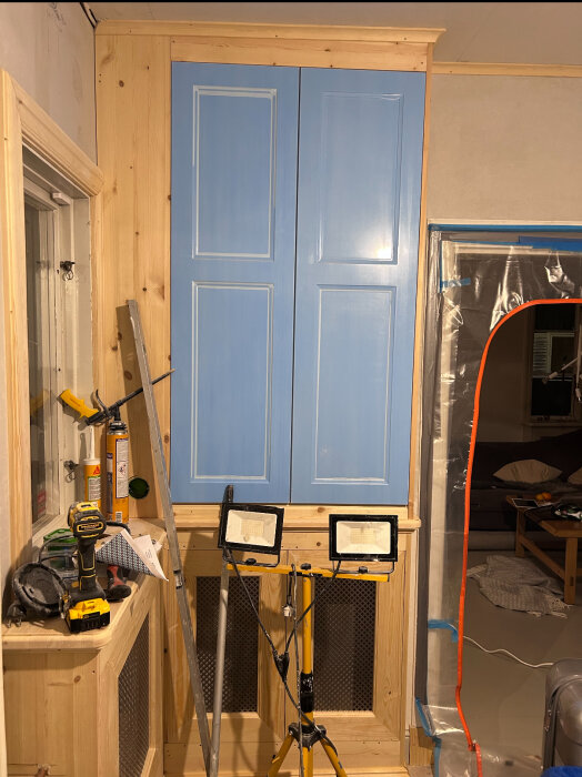 Blå hallmöbel under snett tak, träpaneler syns, bygglampa och verktyg bredvid. Diskussion om att dölja snedheten med färg.