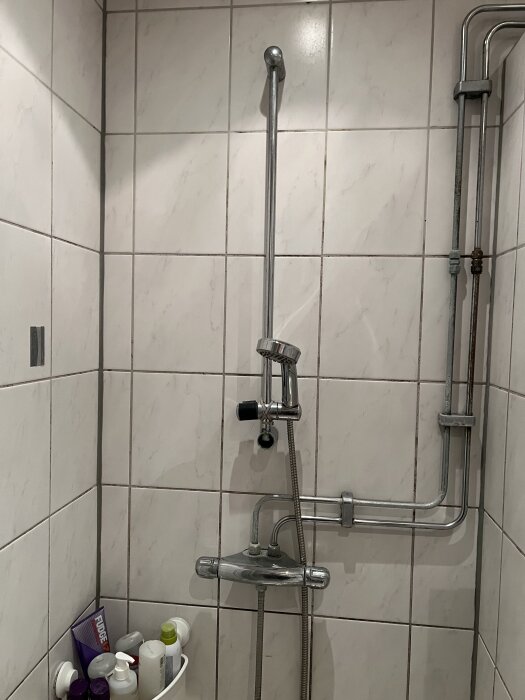 Dusch med silverblandare och rör mot vita kakelväggar; kalkavlagringar kan synas. Flaskor med duschprodukter i ett hörn.