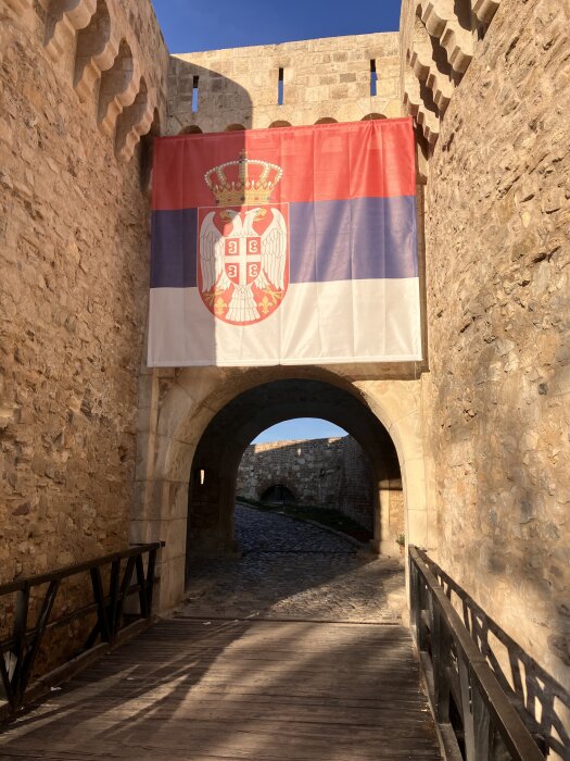 Flagga med rött, blått och vitt hänger över en stenbåge i en gammal befästning med kullerstensväg.