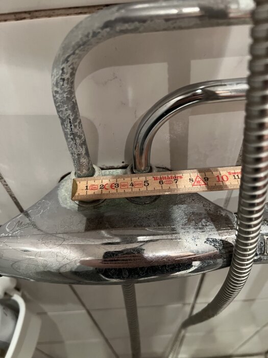 Närbild på en duschblandare med tydliga kalkavlagringar, mätsticka visar mått på 10 cm, kakelplattor i bakgrunden.