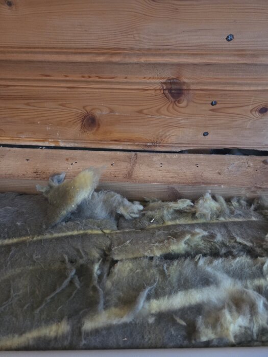 Vägg i trä med synlig isolering och skav vid mötet mellan tak och vägg under renovering, där fuktspärrsplasten behöver ersättas.