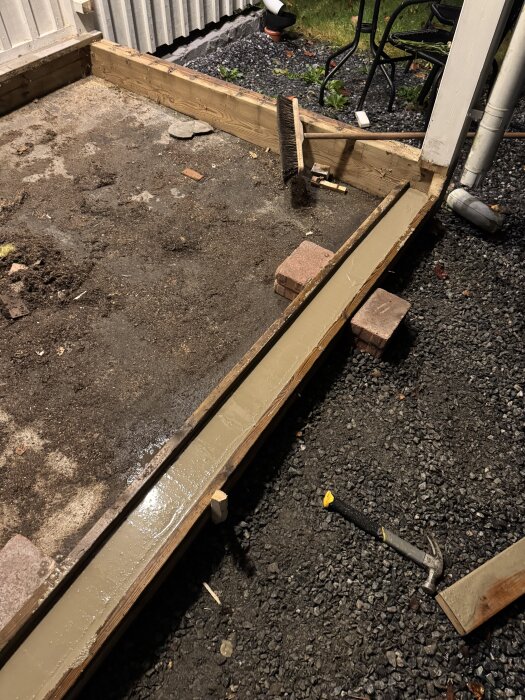 Gjutform med nylagd betong i farstu, omgiven av grus och en hammare. Brädor och en borste ligger vid kanten.