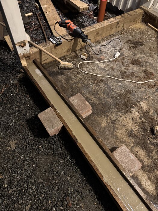 Byggplats för farstu med lutande golvplatta, verktyg som såg och borste på jorden, samt trä- och tegelstödd struktur under bygge.