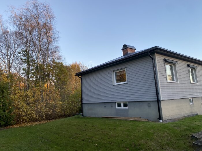 Gaveln av ett hus med träfasad och ett tegeltak, omgivet av en grön gräsmatta och träd i bakgrunden under en klar himmel.