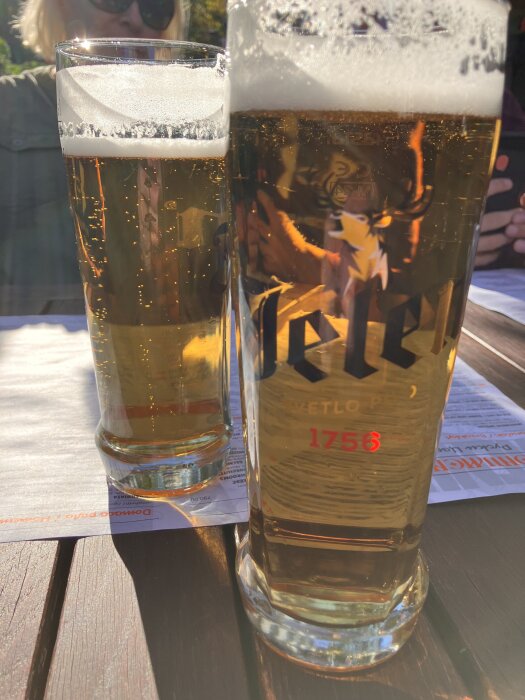 Två glas med öl på ett bord utomhus, med bubblor och skum, soligt väder och en person i bakgrunden.
