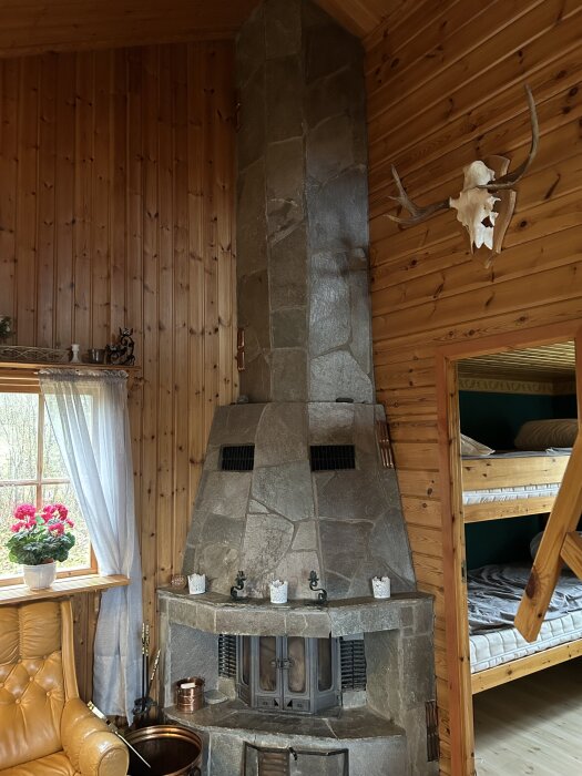 Skifferklädd eldstad i träpanelvägg rum med fönster, krukväxt, och jakttrofé på väggen. Intill finns en våningssäng i trä.