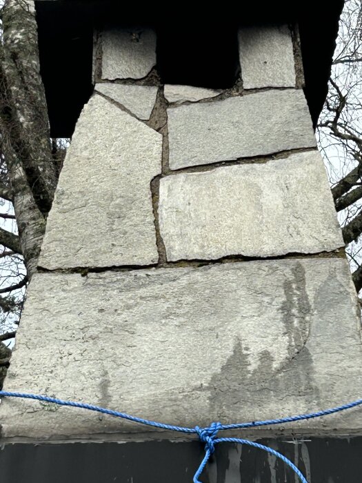 Skorsten i skiffer med sprickor, omgiven av trädgrenar, blått rep vid basen, frågor om rörinsättning utan rivning.