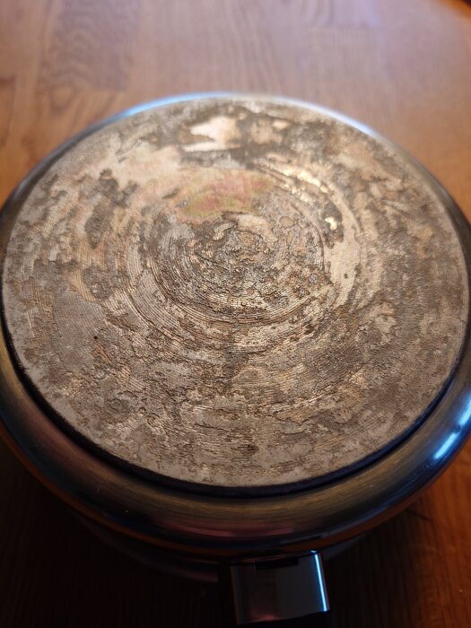 Undersida av kastrull med ojämn beläggning och missfärgningar på en träyta.