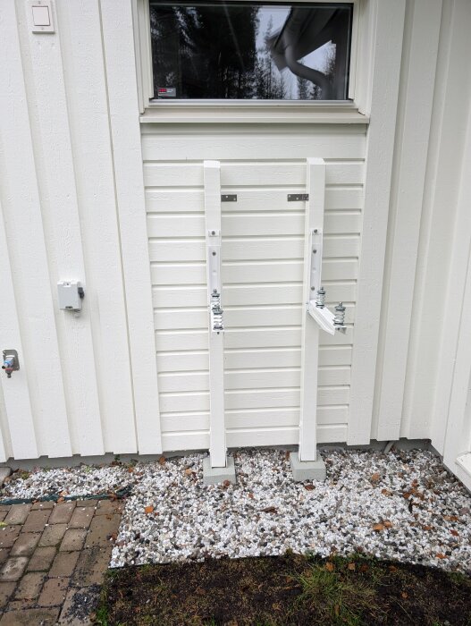 Vita väggfästen monterade på husets träpanel, placerade på små plintar, omgivna av grus och del av stenväg.