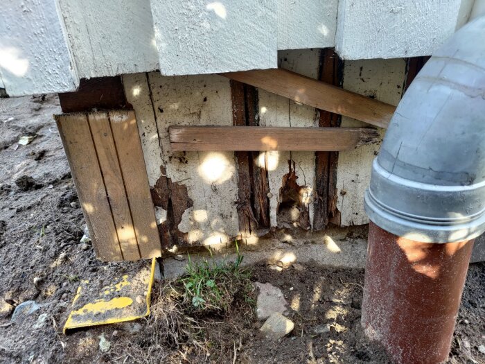 Ruttna träribbor och en rostad metalleternitplatta vid grunden av ett garage, samt ett stuprör och jordig mark i bakgrunden.