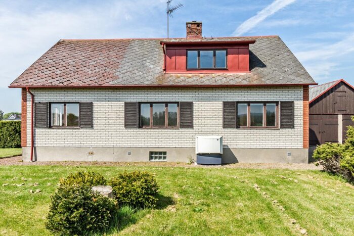 Ett tegelhus med brun- och vitfärgad fasad, två fönsterluckor och en vit Thermia Atria utedel på gräsmattan framför huset.