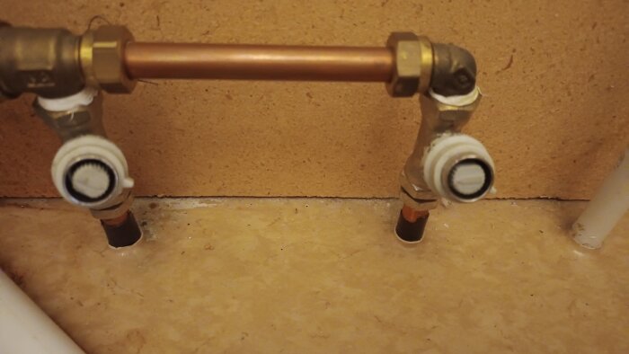 Två rörledningskopplingar under radiator, med vit justeringsventil, anslutna till kopparrör mot en bakgrund av beige vägg och golv.