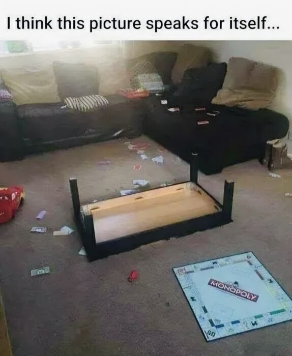 Omkullvält bord i ett vardagsrum med Monopoly-spel och utspridda sedlar på golvet, bredvid en mörk soffa.