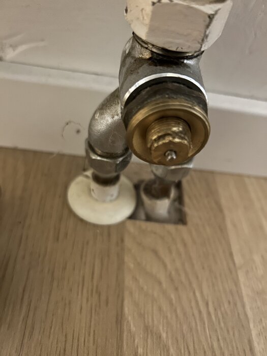 Närbild av en ventilanslutning i metall vid en radiator mot en vit vägg, monterad på ett golv av trä.