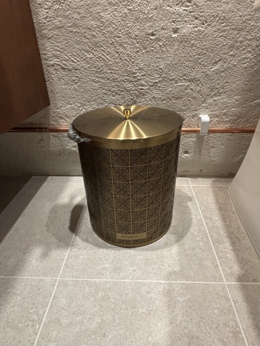 Sopkorg i mässing från Rituals placerad på ett ljust kakelgolv med en strukturerad vägg bakom.