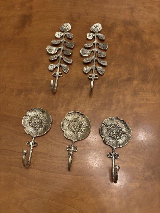 Fyra dekorativa krokar i mässing, två blomstermotiv och två bladmönster, placerade på en träyta.