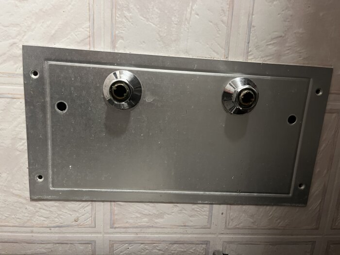 Metallpanel på vägg med två vattenrörsanslutningar för en blandare, omgiven av fyra monteringshål.