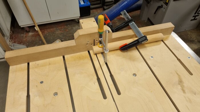 Träsläde med t-spår och vinklade borrhål, används för att hålla runda träbitar med tvingar på en arbetsbänk.