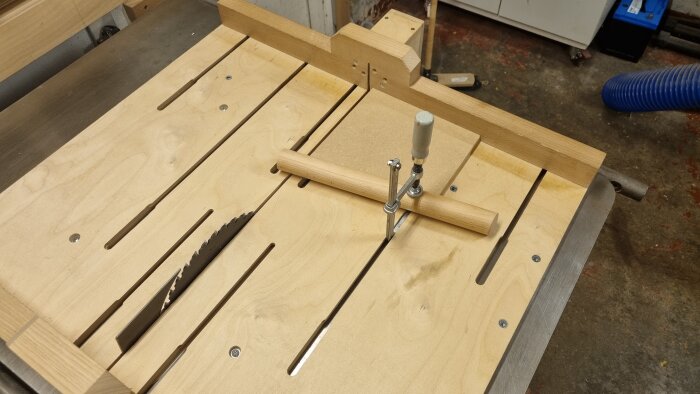 En träarbetssläde med t-track och klämma monterad på ett sågverktyg för att hålla runda ämnen stadigt vid sågning.