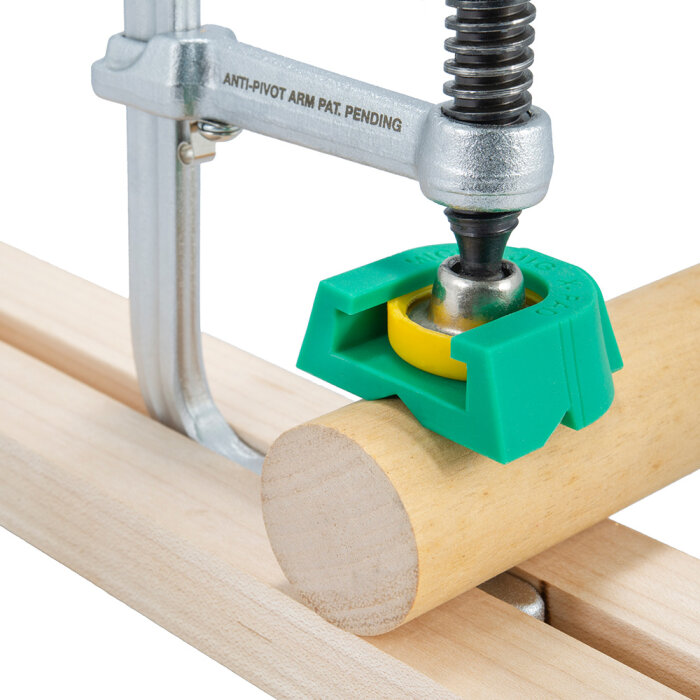 Verktygsklämma med anti-pivot arm håller en rund trästång fast mot en yta med en grön och gul hållare för att stabilisera vid sågning.