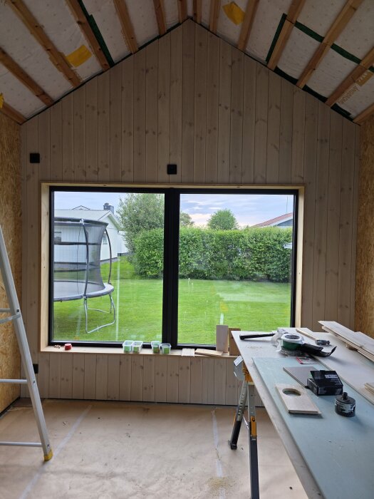 Rum under renovering med nyspikad innerpanel utan lister. Ett fönster visar en trädgård med studsmatta. Verktyg och material på ett bord.