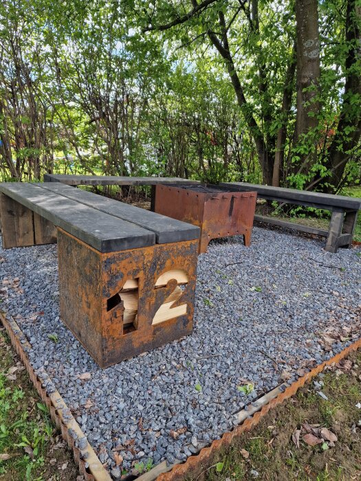 Grillplats med rostig metalldesign, omgiven av träbänkar på en yta med grus och träd i bakgrunden.