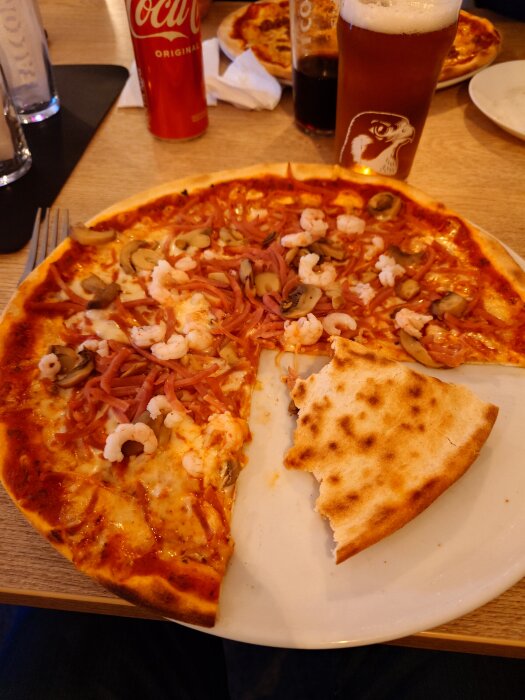 Pizza med räkor, skinka och svamp på en tallrik, omgiven av drickglas och en colaburk på en restaurang.