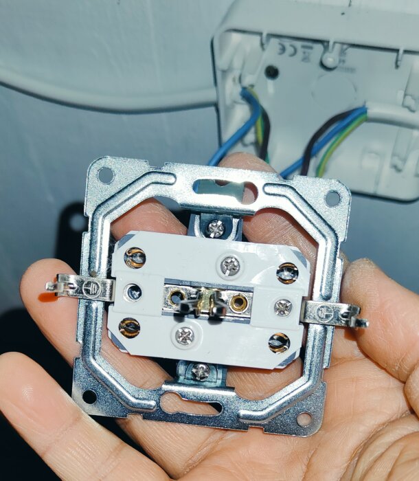 En hand håller en väggmonterad eluttagstomme med synliga skruvar och kabelanslutningar. Bakgrunden visar en väggdosa med anslutna kablar.