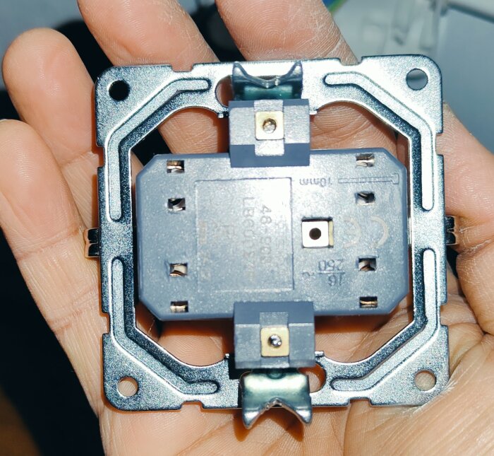 Ett vägguttag utan täckplatta hålls i en hand, med synliga skruvhål och metallram.