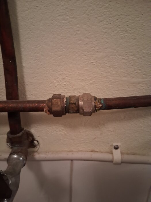 Gammalt kopparrör med klämringskoppling och synliga tecken på korrosion i en källare, anslutet till ett duschblandarsystem.