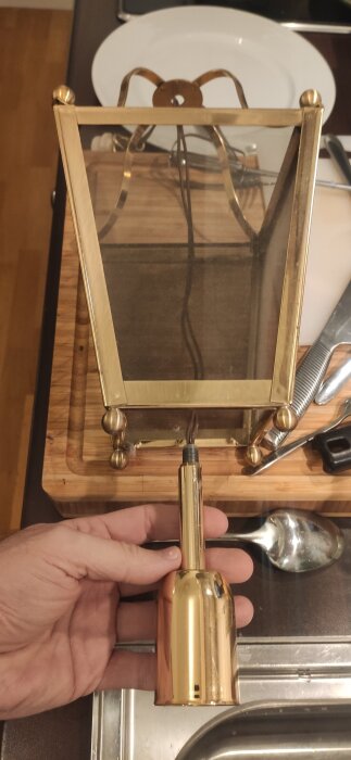 Guldfärgad lampa med glasruta, metallstativ och synlig kabel, undersöks för jordningsmöjligheter. En hand håller lampans bas på en köksbänk.