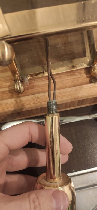 Hand håller en armaturdel med två elkablar, sett nära ett metallrör. En räfflad mutter syns på röret.