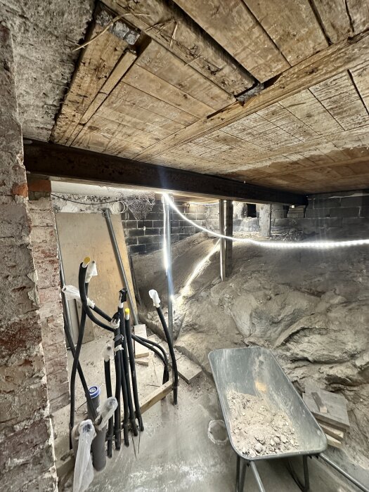 Källarutrymme under renovering med synlig stålbalk i taket, jord och berggrund, samt en skottkärra fylld med grus och byggmaterial.