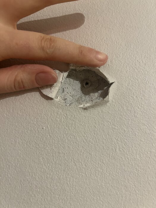 Hål i vägg där betong bakom ytskikt syns, sprucken med lösa bitar; finger pekar för att visa skadans omfattning.