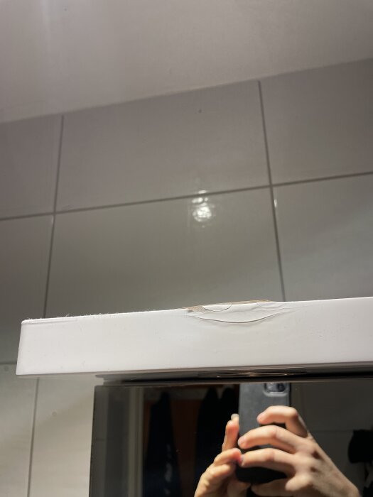 Skada på ytan av en vit möbel på grund av läckande vattenpistol, reflektioner i spegel, händer håller en mobilkamera.