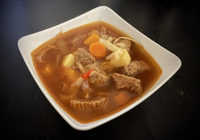 Hemgjord köttsoppa med kalvkött, morötter, potatis, butternutpumpa, lök och paprika i röd buljong serverad i en fyrkantig vit skål.
