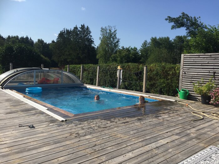 Utomhuspool med trätrall runtom, två barn badar. Soligt väder och grönskande häckar i bakgrunden.