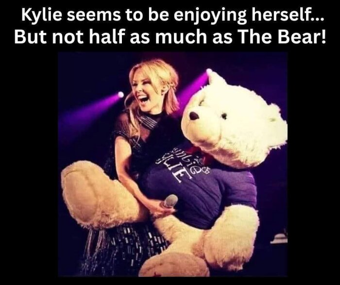 En kvinna ler och håller en stor nallebjörn på scenen, med text ovanför dem.