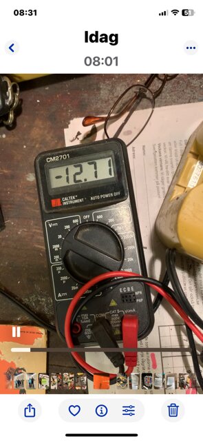Multimeter visar -12,71 volt bredvid taget Topcon Laser-batteri, med ledningar anslutna och delar av verktygsbordet synligt.