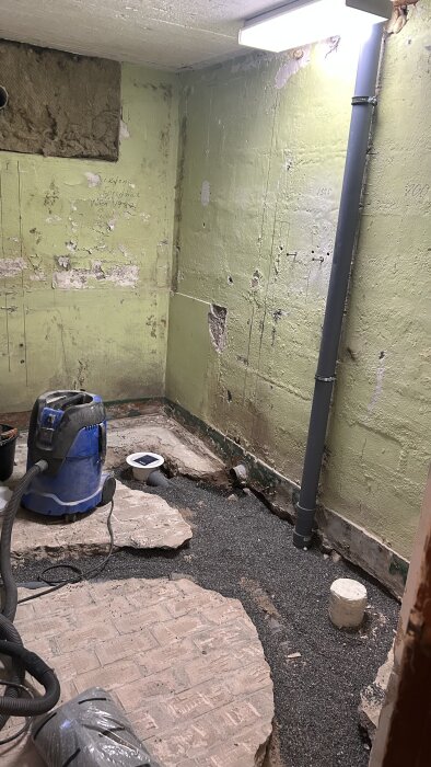 Badrum under renovering i källare med gröna, ojämna väggar och delvis uppbrutet golv, dammsugare och synliga rör.